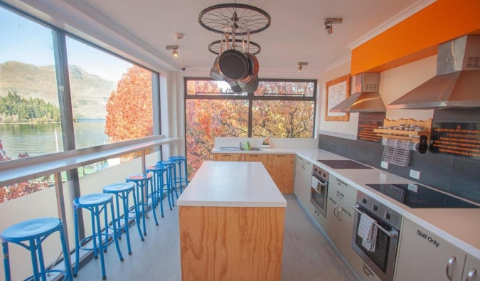 Sunny and expansive kitchen with stoves, island, and line of stools by a window-side counter overlooking the water and mountains at Absoloot Hostel in Queenstown, New Zealand