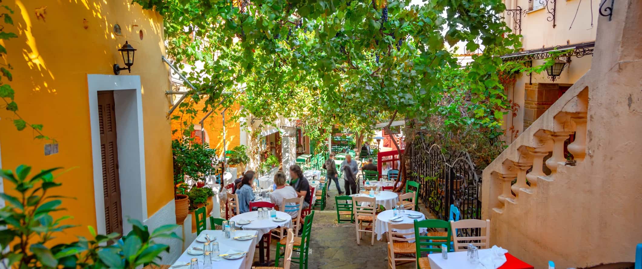 Cafe tables lining an alleyway covered with a lush tree branches, surrounded by bright yellow buildings, in Athens, Greece.