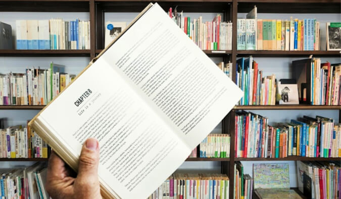 Hand holding a book open at Chapter 8 in the background are shelves full of books